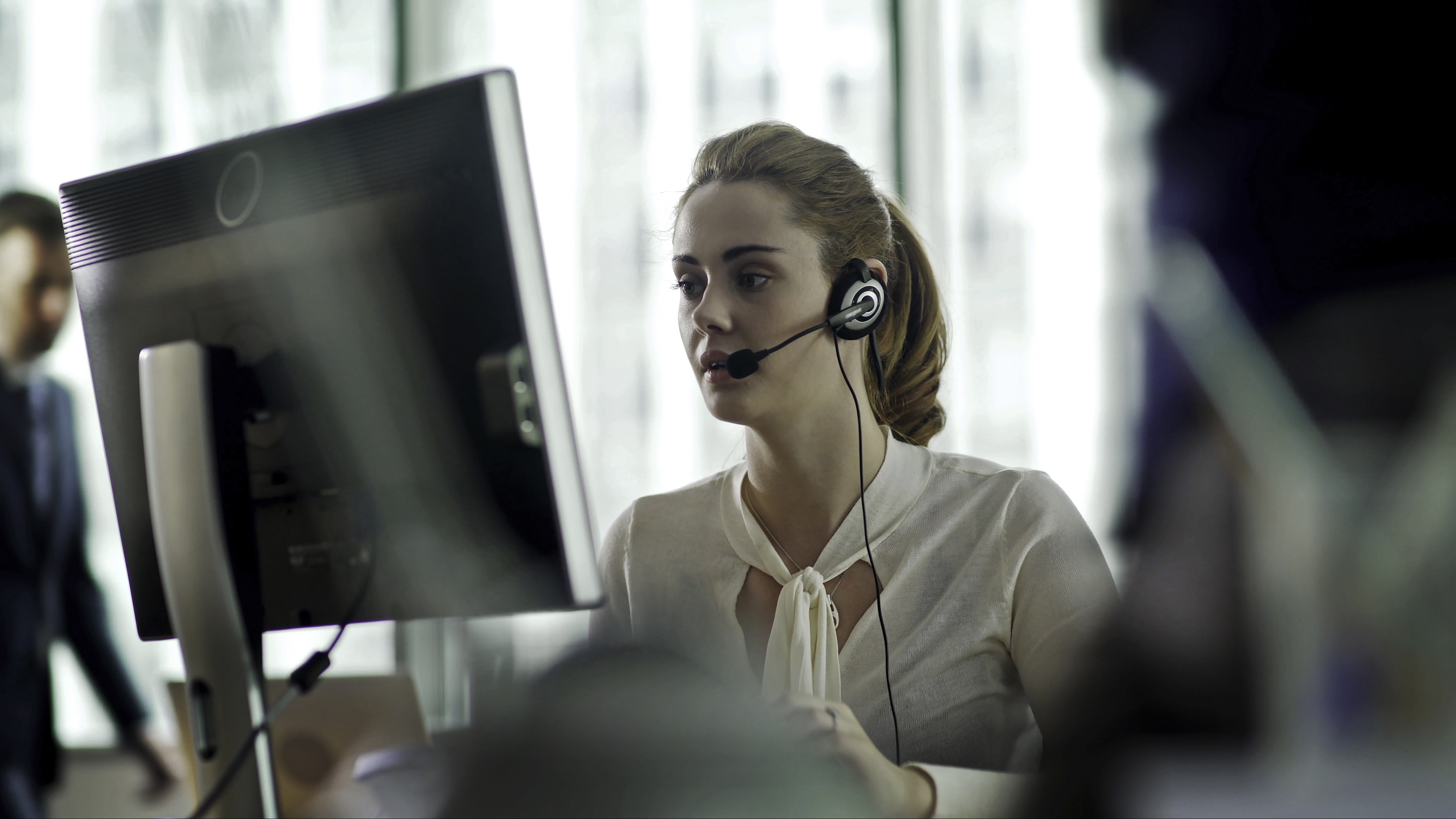 A good looking woman sitting at a desk, surrounded by her co-workers, communicating with someone using a headset &amp; referring to her computer display while she talks on the phone.